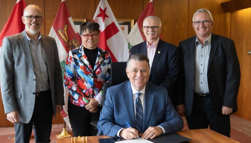 image of four people standing behind and overlooking a person while someone sits at the desk signing a proclamation