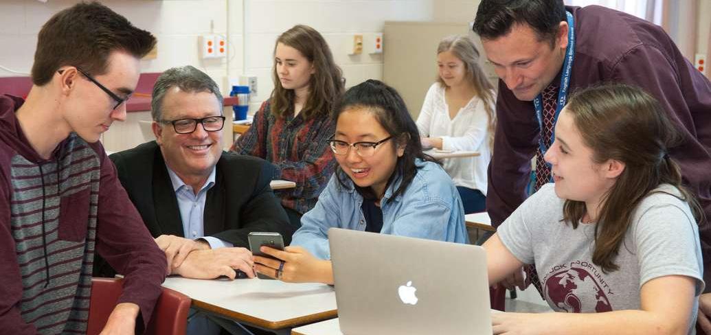 Minister Currie, a teacher and three high school students looking at a smart phone and a cumputer.