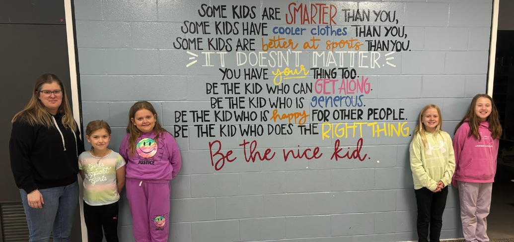 image of a teacher and elementary students standing near a wall with some inspirational writing on it.