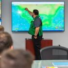 In the emergency operation centre, one man standing and pointing at a screen while more people look on from seats