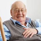 An elderly man with a cheerful expression sits comfortably in a chair, holding a cane in his right hand,